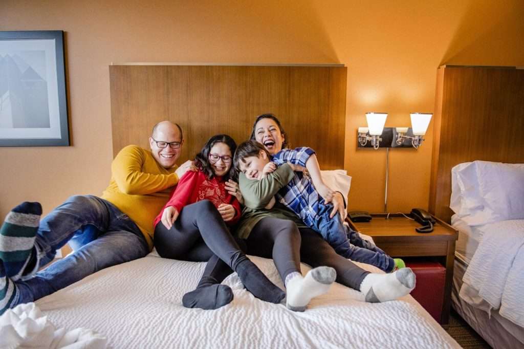 Family relaxing together on a bed in a Halifax hotel room, with warm décor and a second bed nearby.